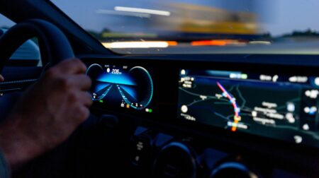The dashboard of a car at night-time, with symbols and displays lit up and the driver's hands on the steering wheel, with blurred external lights in the background