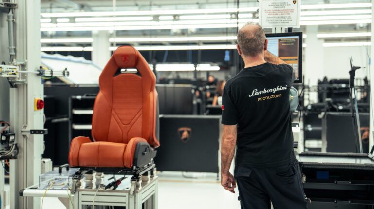 A Lamborghini worker uses a touchscreen display in a warehouse with an orange vehicle seat to his left under construction