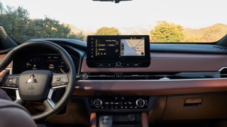 Inside the Mitsubishi Outlander 2026. The dash is predominantly brown, with a VDU next to the steering wheel . The tops of trees are visible through the windshield.