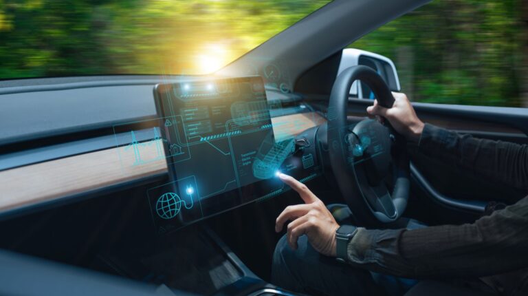 A driver sits at the wheel of a connected SDV, with lights on the display and wheel depicting its technology. A green background is visible through the windshield and driver window.