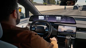 Interior view of the driver's position in the Luci Gravity, with a wide touchscreen display, steering wheel and a quiet highway scene visible through the windshield.